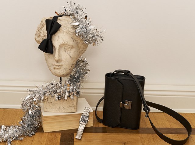 Klassisk byste prydet med en sort sløyfe og sølvfrynse, hvile på en bok med et sølvur ved siden av, ved siden av en svart håndveske på tregulvet.