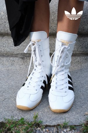 High-top sneakers in white leather with black stripes, featuring a gum sole and gold accents. Laces are white, with a padded collar.
