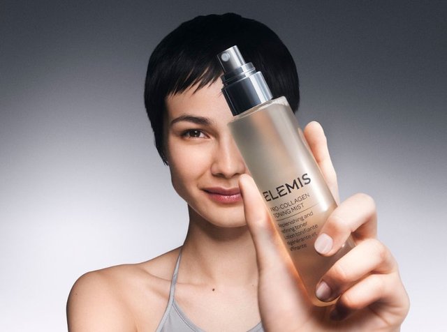 Przezroczysta szklana butelka ELEMIS Pro-Collagen Toning Mist; metaliczna srebrna nakrętka w sprayu, lekki płyn, z etykietą, trzymana przez osobę z krótkimi czarnymi włosami.