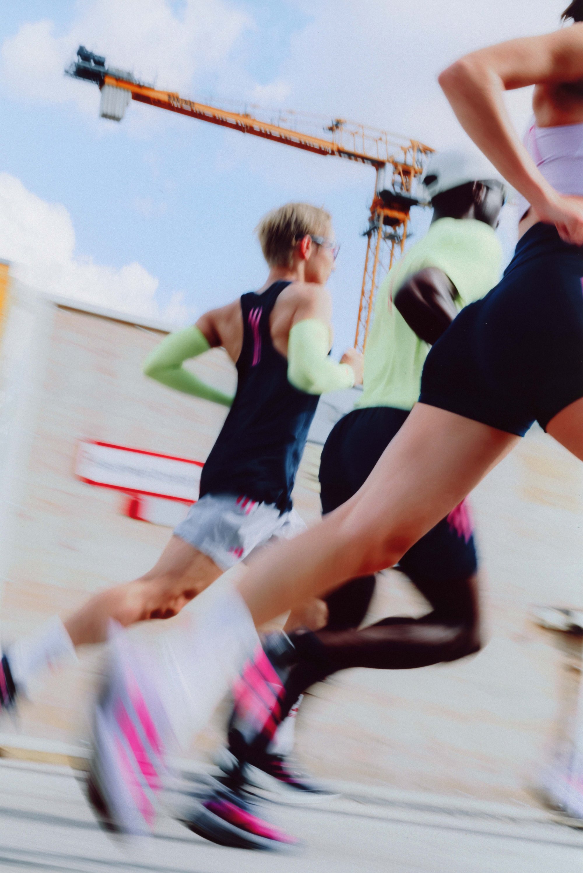 A group of runners in motion. One is wearing a black tank top with green sleeves, while another is dressed in yellow. Their shoes and shorts feature distinctive pink accents.