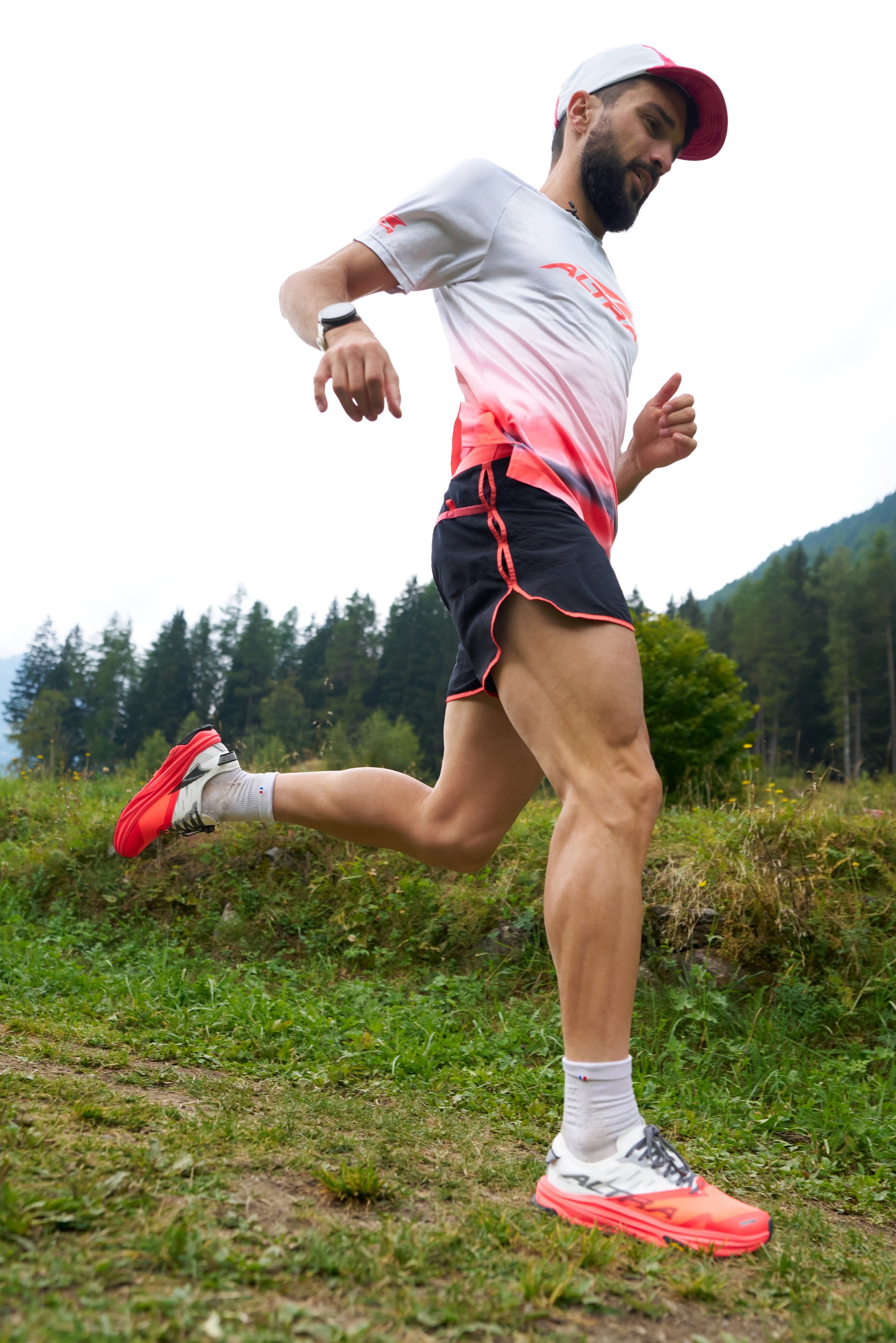 Runner wearing a red and white gradient shirt and black shorts. Bright red shoes with grey accents and white socks, standing on grass with trees in the background.