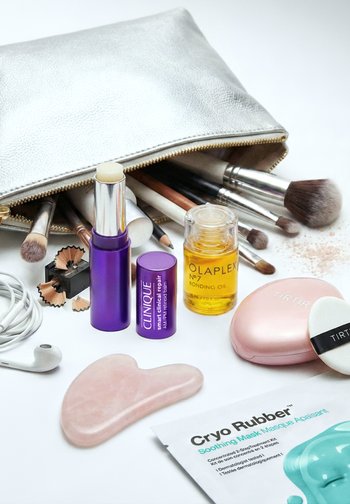Borsa trucco argento con pennelli, balsamo labbra Clinique, olio ristrutturante Olaplex, cipria compatta Tirtir con applicatore, gua sha in quarzo rosa, maschera viso Cryo Rubber.
