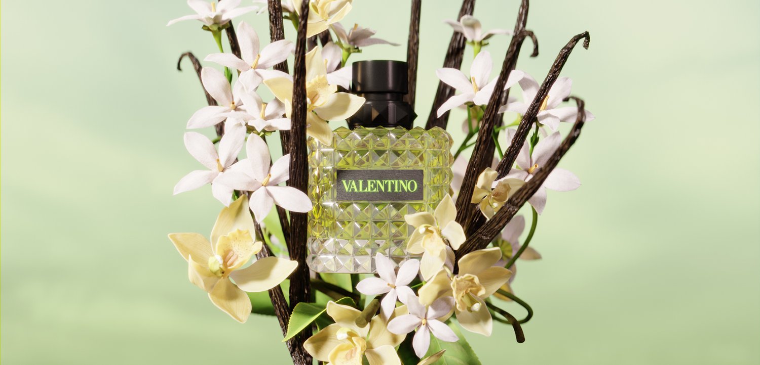 Parfumflesje met label Valentino, omringd door witte en gele bloemen en donkerbruine vanillepeulen tegen een zachte groene achtergrond.