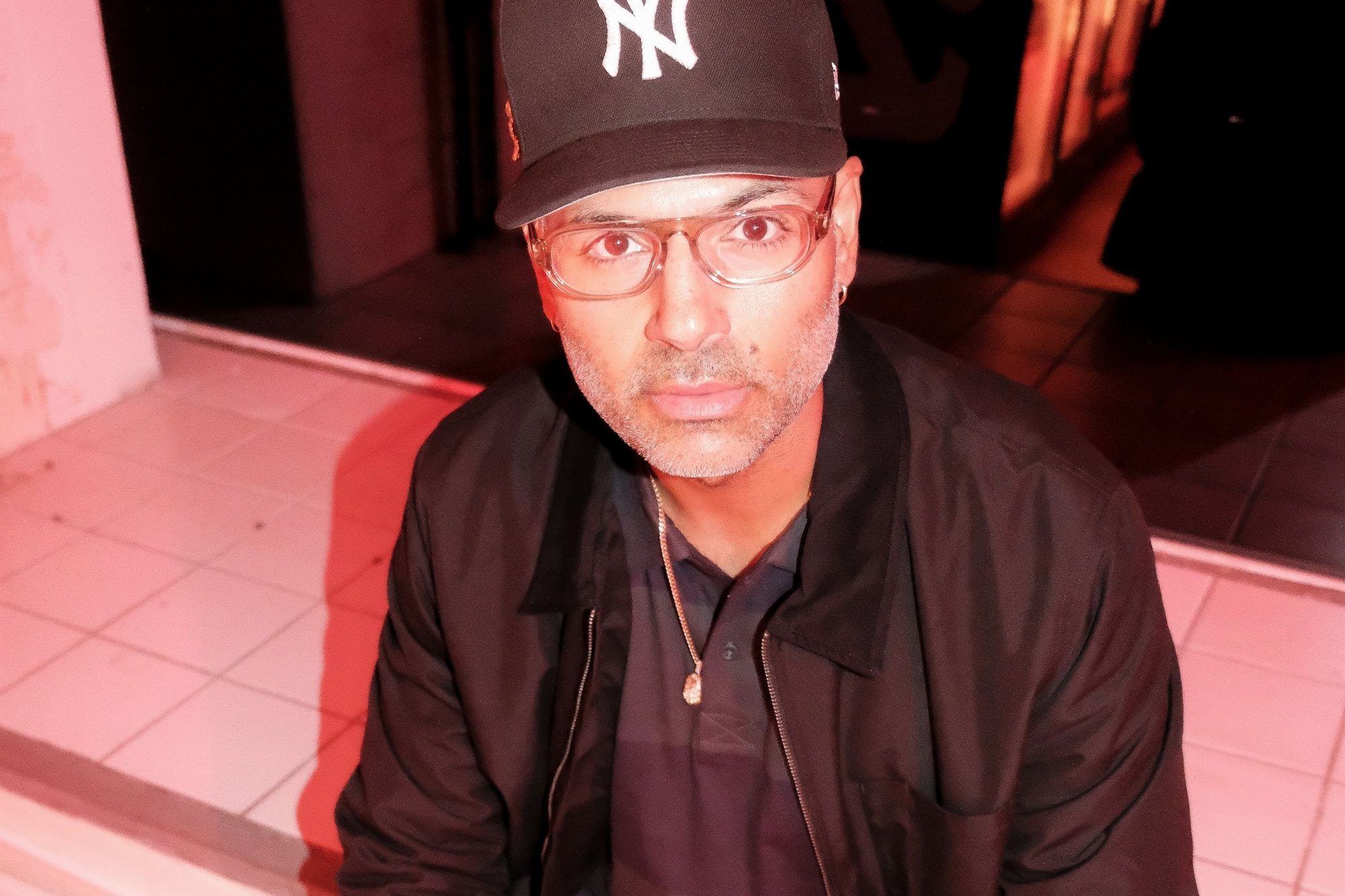Black baseball cap with white logo, clear glasses, black jacket over dark shirt, sitting on white tiled surface with red lighting.