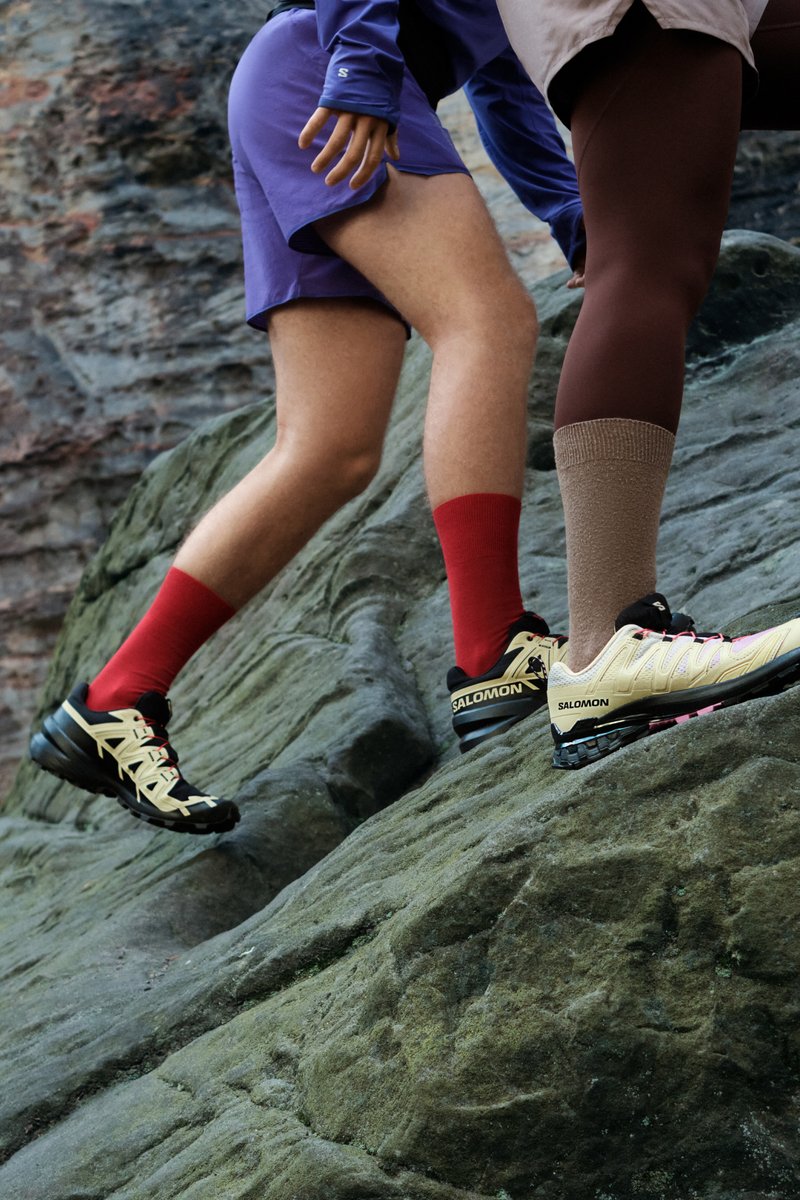 Scarpe da trail nere e crema con dettagli gialli. Calzini rossi e marroni. Terreno roccioso con una superficie texture sullo sfondo.
