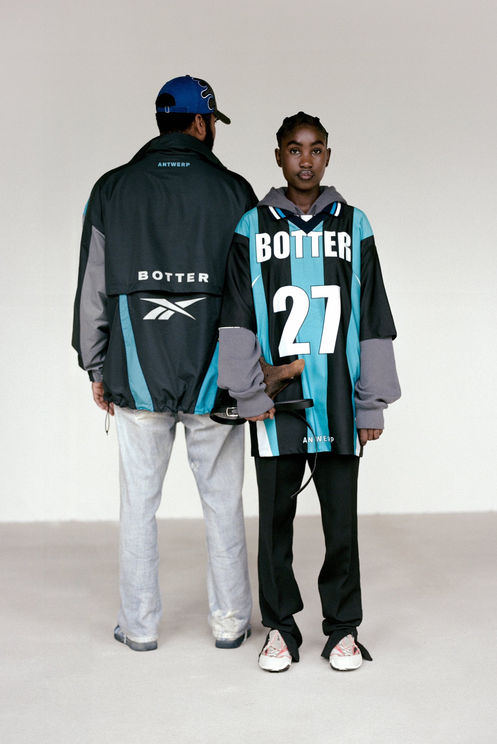 Un hombre con una chaqueta negra y teal se encuentra detrás de una chica que lleva una camiseta teal de gran tamaño con 'BOTTER 27'. Ambos están en un entorno minimalista.
