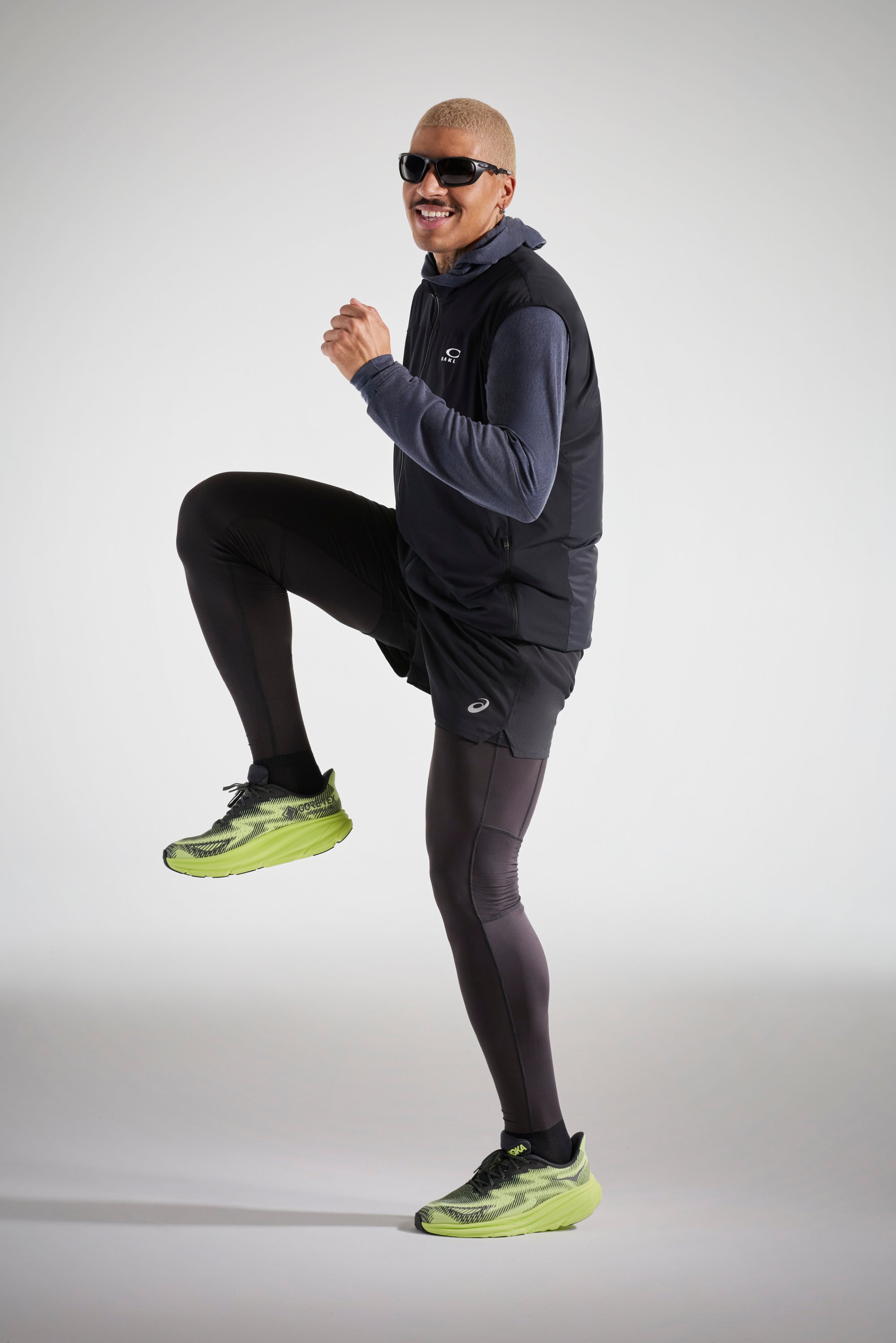 Una persona sonriente con cabello rubio lleva gafas de sol, un chaleco sin mangas y leggins deportivos, capturada en medio de un ejercicio, levantando una rodilla.