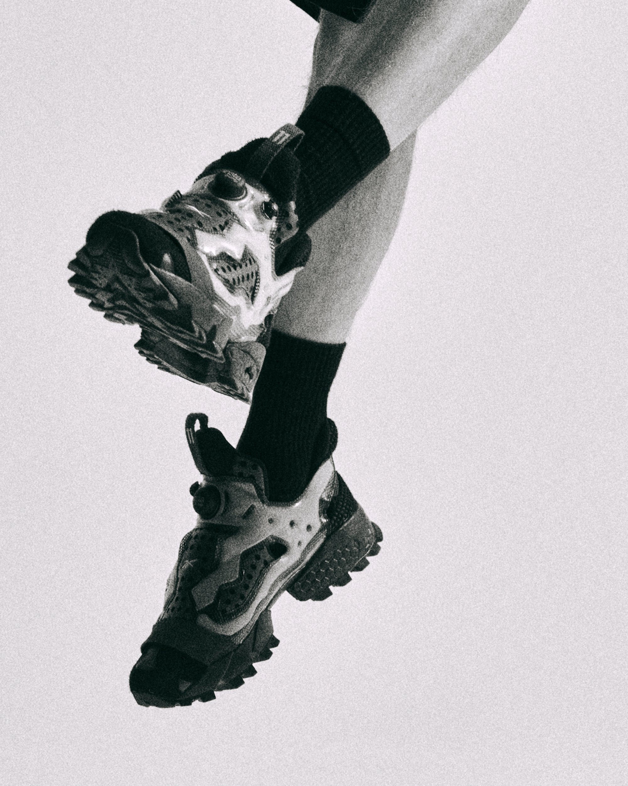 Se muestran zapatos con un diseño único, llevados con calcetines oscuros; la pierna está posicionada de manera dinámica sobre un fondo claro.