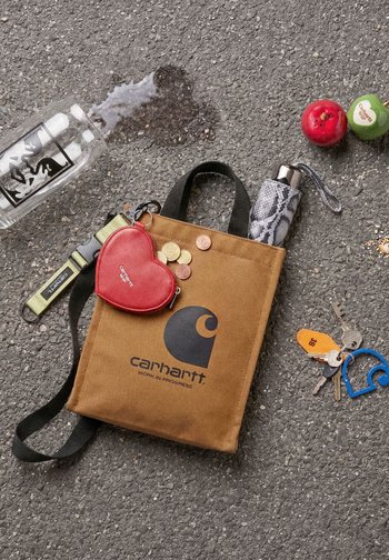 Bolsa marrón Carhartt con monedas, monedero rojo en forma de corazón, paraguas, llaves, dos objetos con forma de fruta y botella de agua derramada sobre el asfalto.