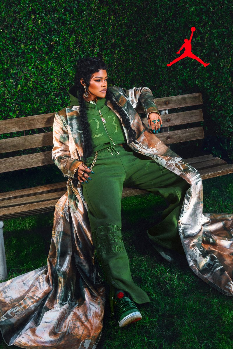 Person with long braids wearing green hoodie and pants, patterned long coat, sitting on wooden bench against leafy background.