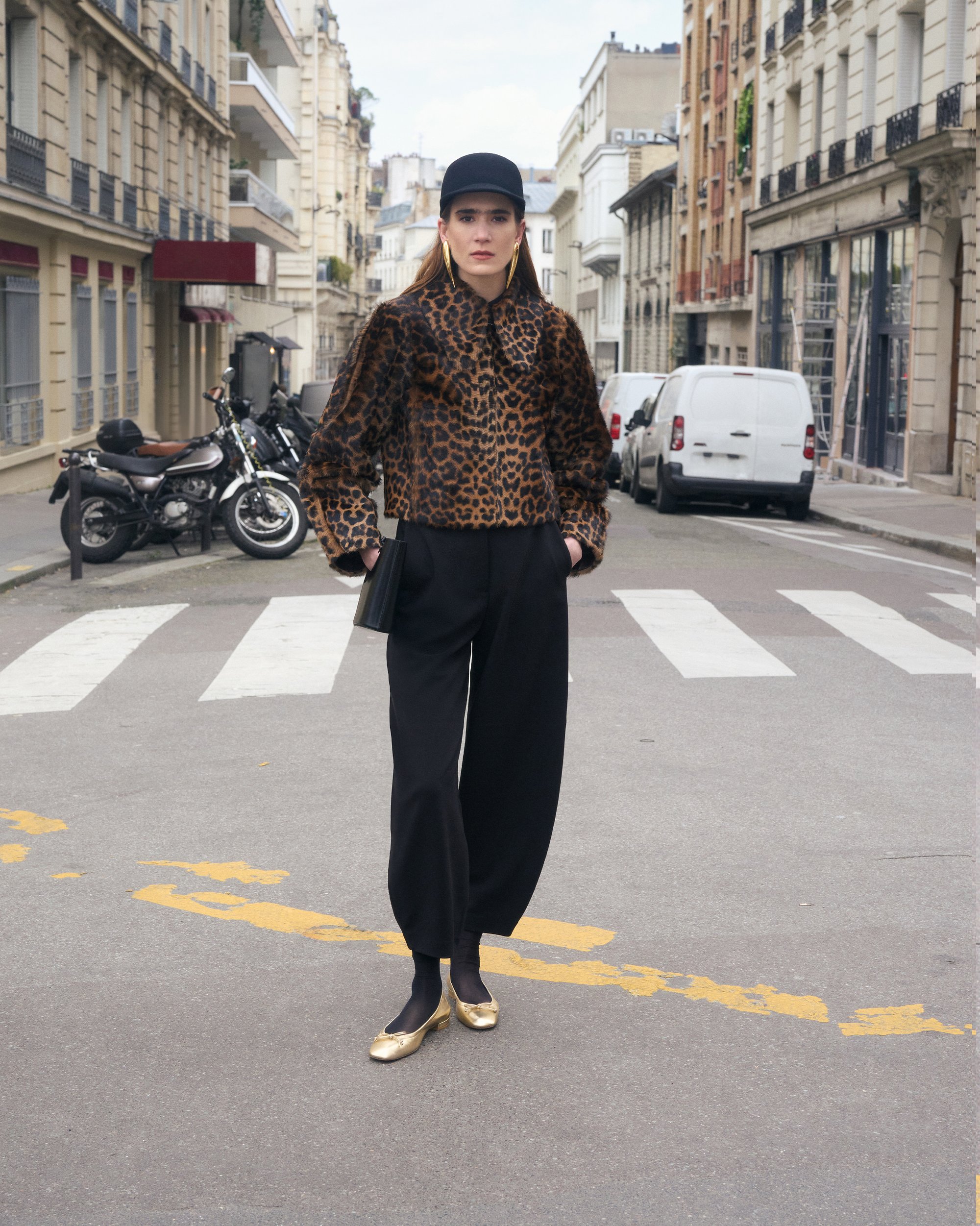Una modelo que lleva una chaqueta de estampado de leopardo, pantalones negros y zapatos planos dorados está de pie en la calle, flanqueada por vehículos estacionados y edificios.