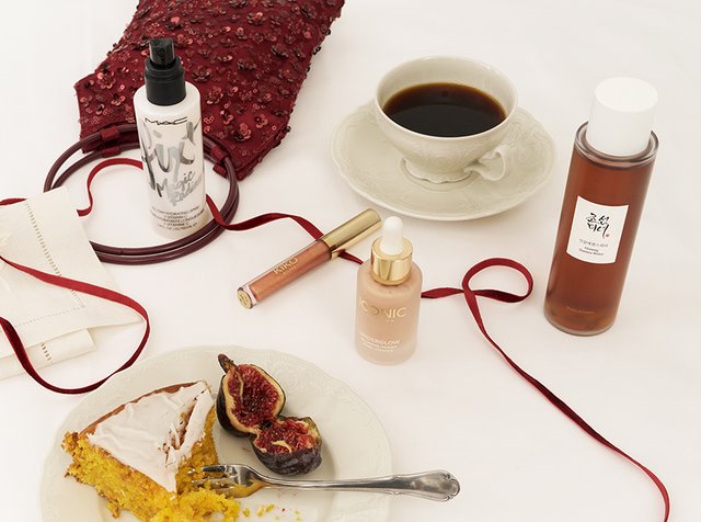 Tarjotin, jossa on kuppi mustaa kahvia, pala kakkua, halkaistuja viikunoita, ihonhoitotuotteita, meikkituotteita ja punainen helmisalkku valkoisella pöytäliinalla.