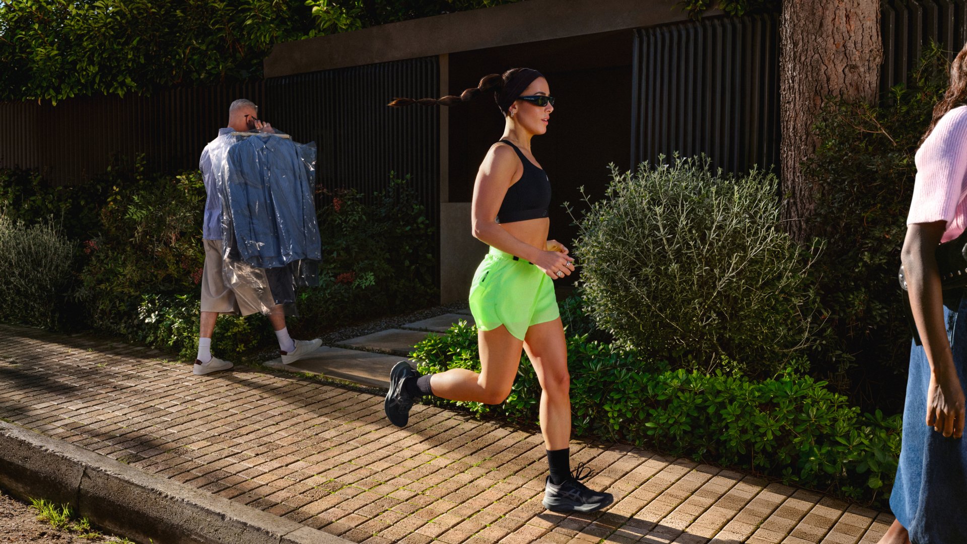 Femme portant un short vert néon et une tenue de sport noire en train de faire du jogging sur un trottoir ensoleillé, tandis qu'un homme marche en portant des vêtements en arrière-plan.