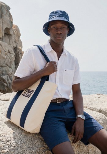 Homme portant un chapeau seau marine, une chemise blanche à manches courtes et un short marine, assis sur des rochers au bord de la mer, tenant un sac fourre-tout beige avec des rayures marines.