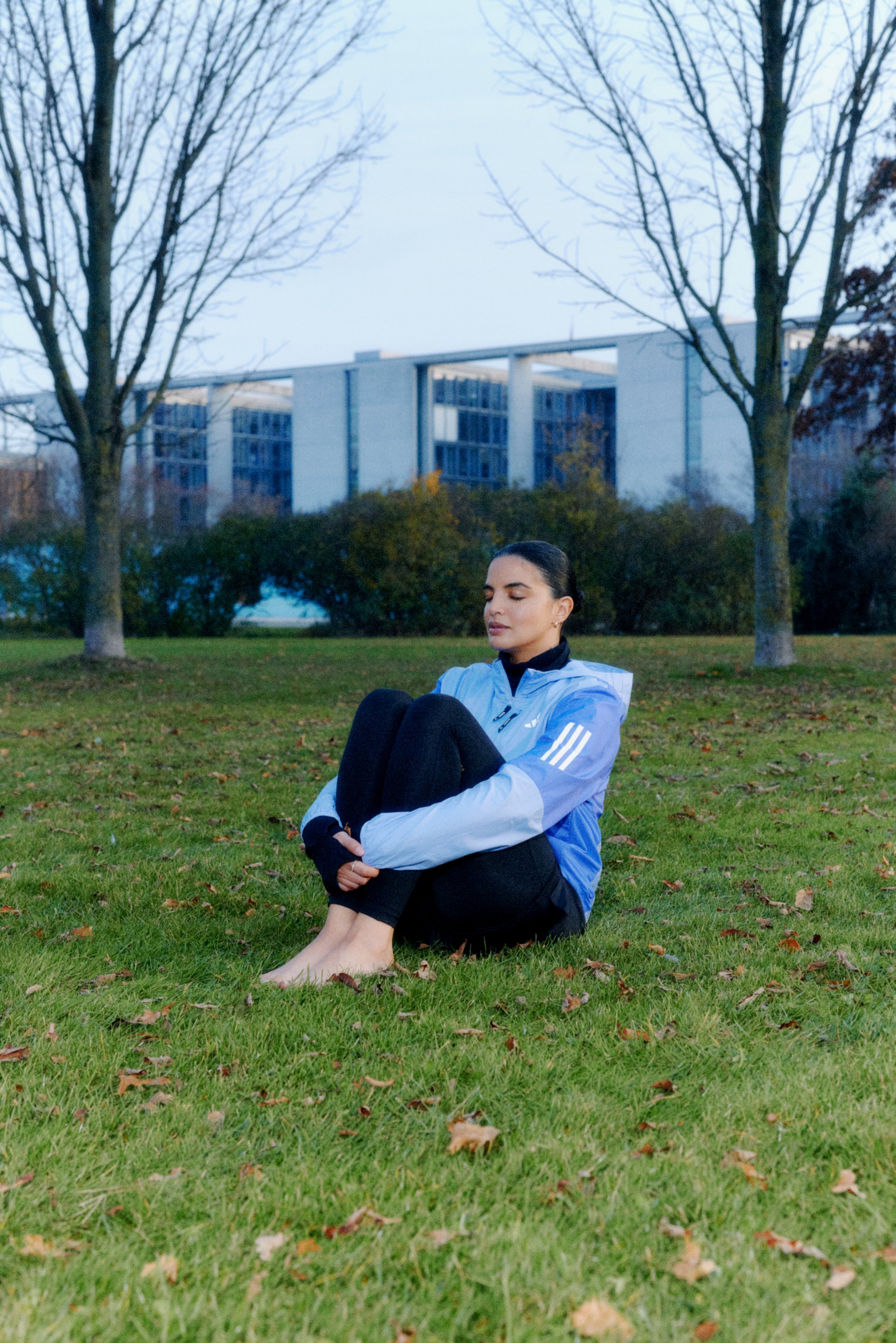 A woman sitting cross-legged on grass, wearing a blue jacket and black leggings, eyes closed, in a serene outdoor setting with trees.