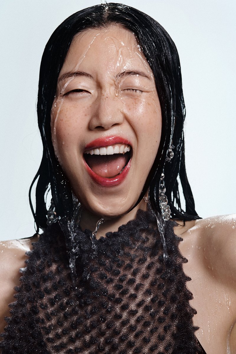 Una mujer con el pelo mojado ríe alegremente, con el agua salpicando su cara. Lleva una blusa negra con textura y grandes pendientes.