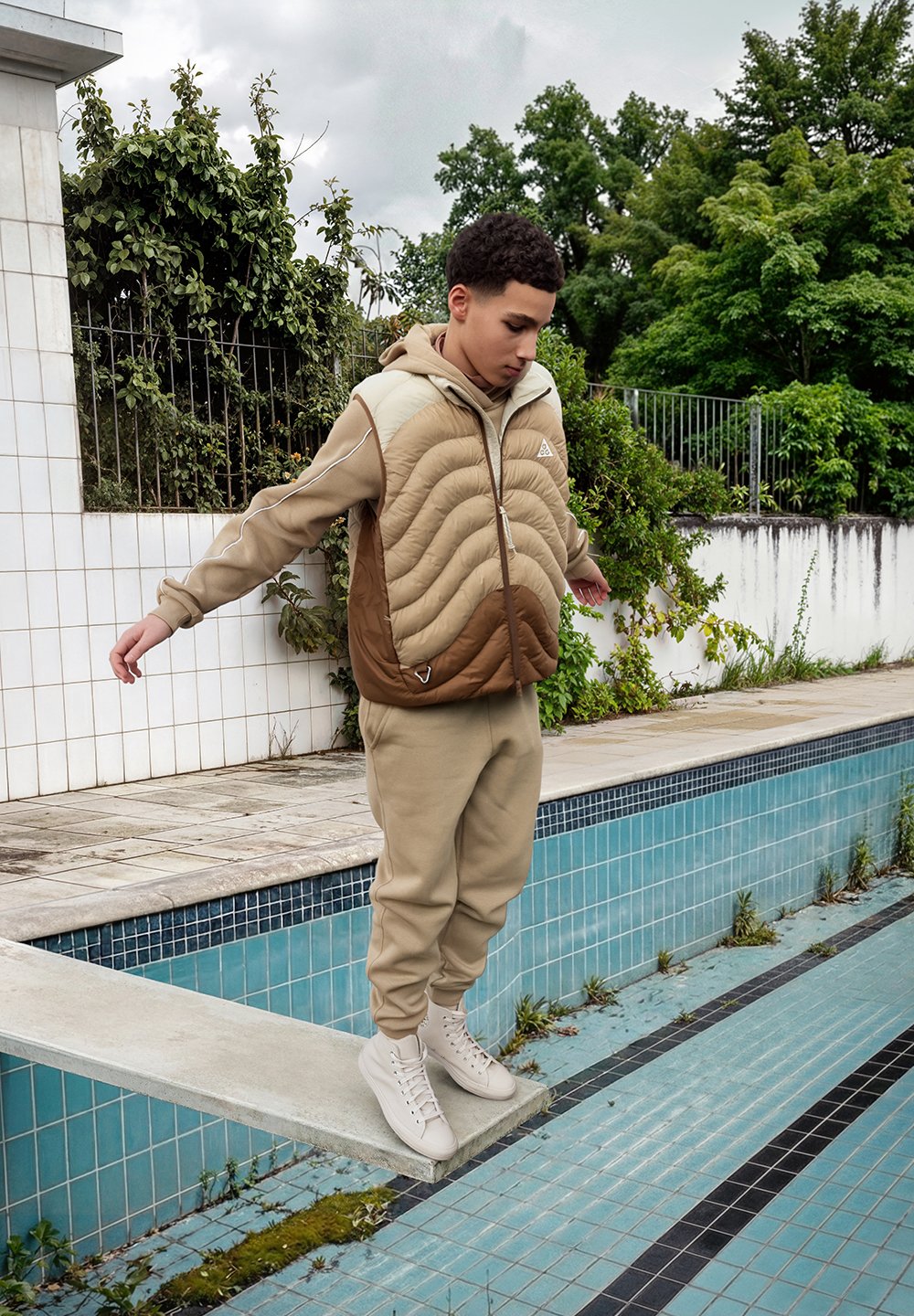 Veste matelassée beige avec motif ondulé, accents marron, pantalon de survêtement beige clair et baskets montantes crème. Debout près d'une piscine. Feuillage vert en arrière-plan.