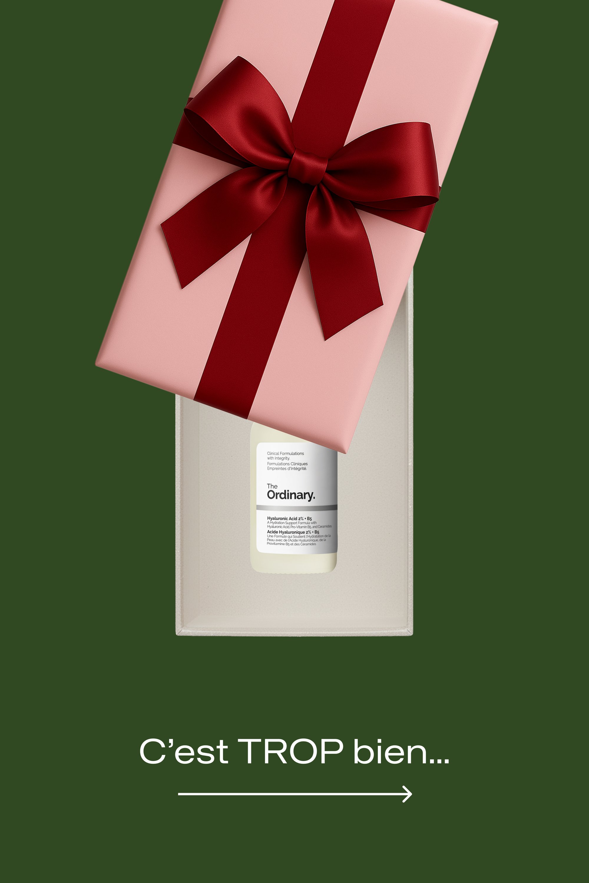 Boîte cadeau en rose pâle avec un ruban rouge brillant, dévoilant une bouteille blanche étiquetée « The Ordinary » avec un texte noir, à l'intérieur d'une boîte de couleur crème.