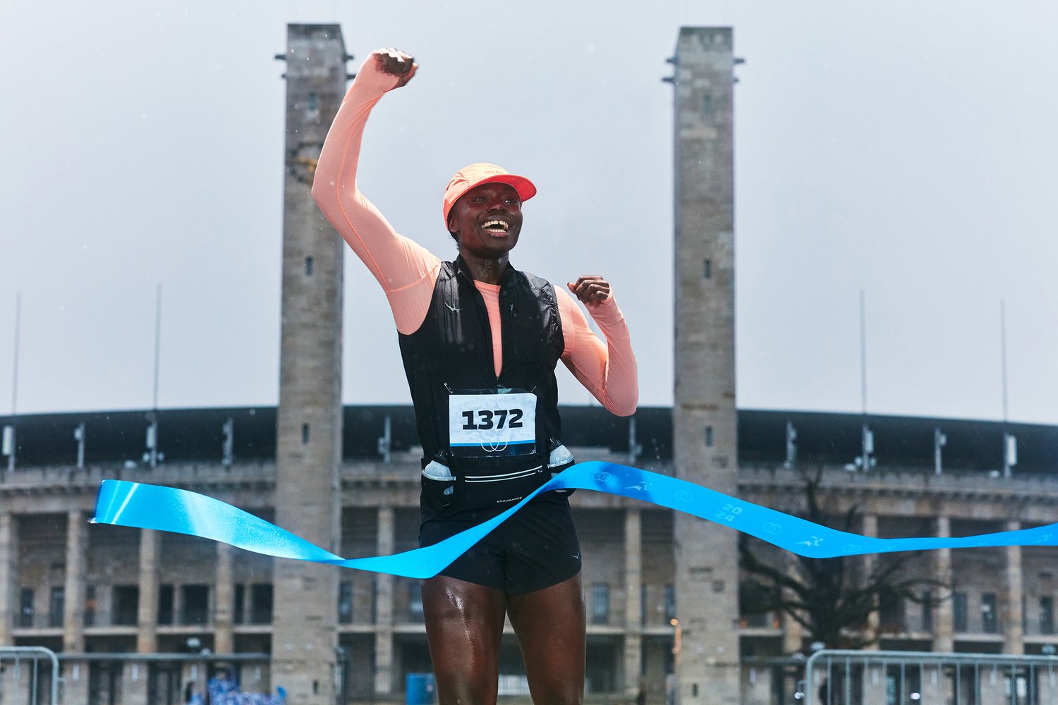 Un atleta alegre con una gorra y camiseta naranjas levanta un puño, rompiendo la cinta de la meta en una carrera al aire libre, con un estadio de fondo.