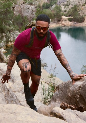 Man met tatoeages en zonnebril die rotsachtig terrein bij een meer beklimt, draagt een rood shirt en zwarte korte broek, met een rugzak.
