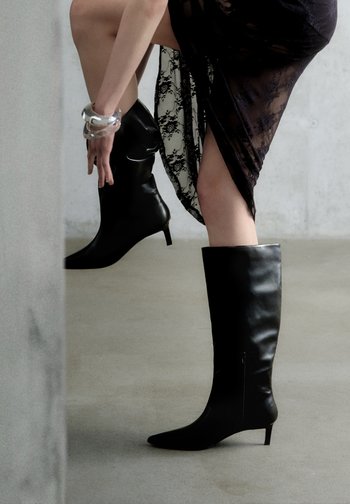 Cizme negre din piele, până la genunchi, cu vârf ascuțit și toc mic. Textură netedă, cu detaliu fermoar lateral. Purtați cu o rochie neagră din dantelă.
