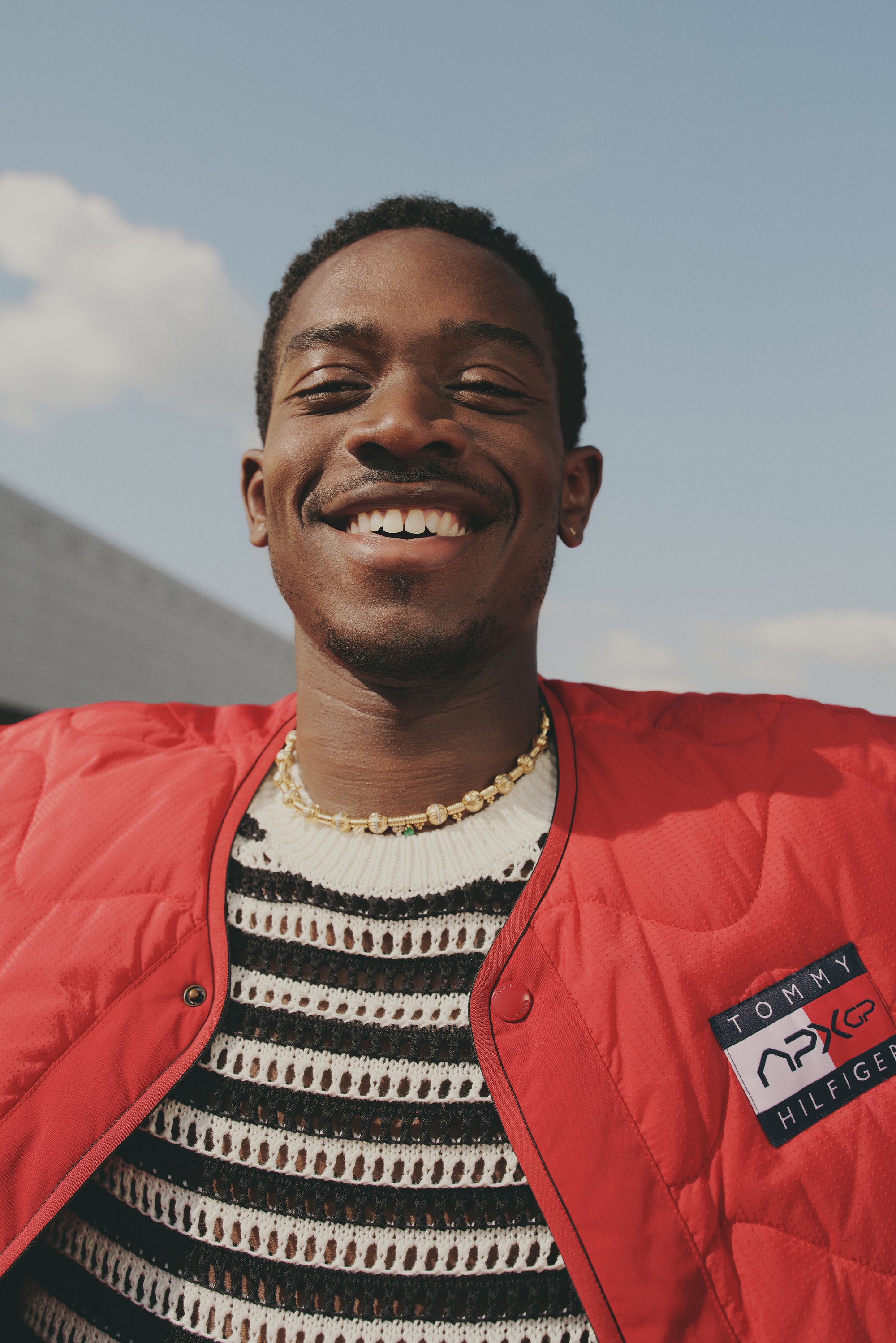 Hombre sonriente con una chaqueta roja con un parche de Tommy Hilfiger, vistiendo un suéter a rayas blancas y negras y un collar decorativo, fondo de cielo azul.