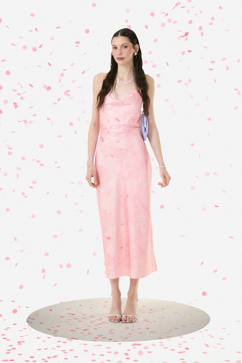 Mujer con el pelo largo y oscuro que lleva un vestido de satén rosa, con un bolso lavanda, de pie sobre una plataforma circular cubierta de confeti.