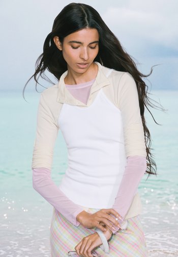 Femme aux longs cheveux foncés se tenant au bord de la mer, vêtue de vêtements pastel superposés, ajustant un bracelet translucide à son poignet.