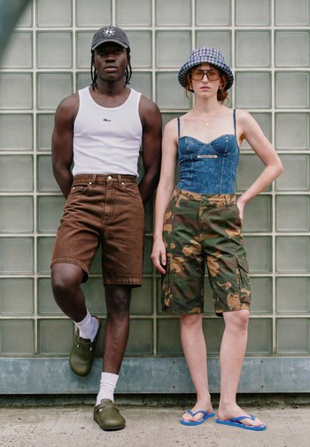 Two young adults standing against a glass block wall, one wearing a white tank top, brown shorts, and cap, the other in denim corset, camo shorts, and hat.
