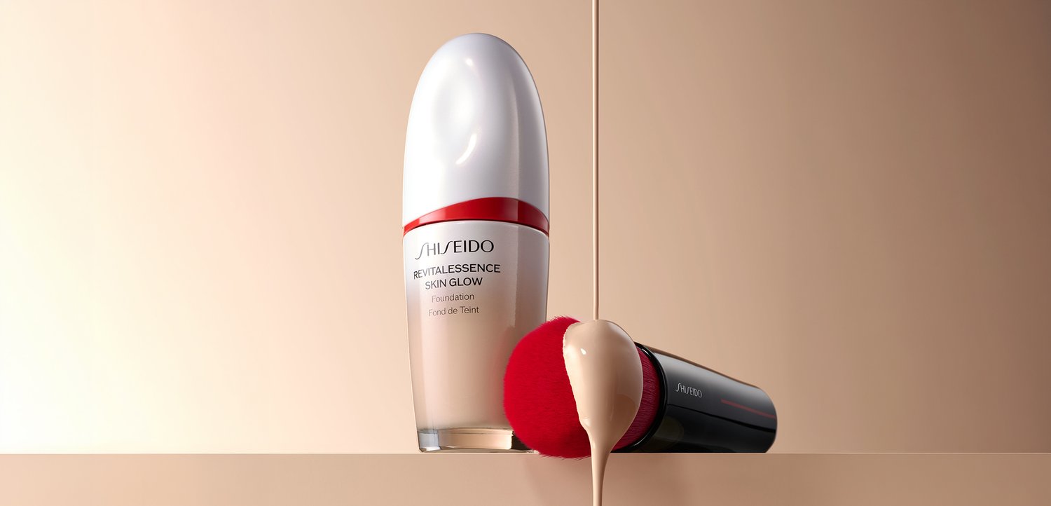 Bottiglia di fondotinta Shiseido Revitalessence Skin Glow accanto a un pennello da trucco con liquido di fondotinta che gocciola su una superficie beige.
