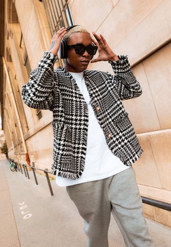 Eine Person mit kurzen blonden Haaren, die eine Sonnenbrille und Kopfhörer trägt, geht entlang einer Stadtstraße in einer gemusterten Jacke und grauen Hosen.