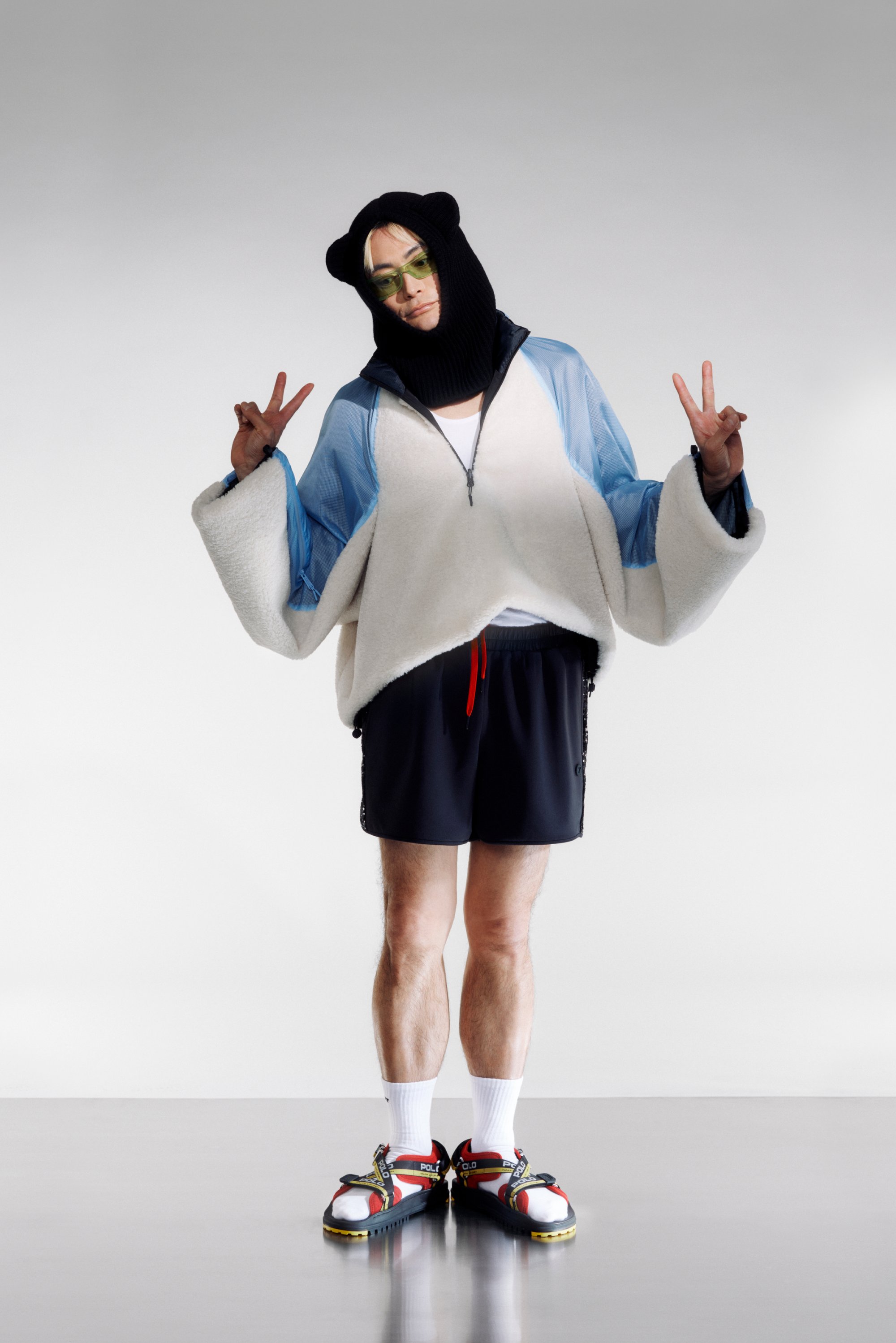 Een persoon staat in een speelse pose, gekleed in een capuchon met berenoren, een blauwe en witte oversized jas, zwarte shorts en kleurrijke sneakers tegen een grijze achtergrond.