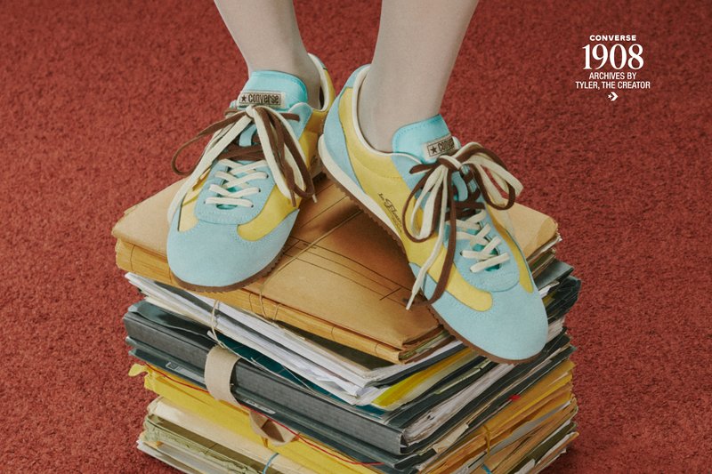 Pieds portant des baskets Converse bleues et jaunes debout sur une haute pile de dossiers et papiers usés et attachés posés sur un tapis rouge.