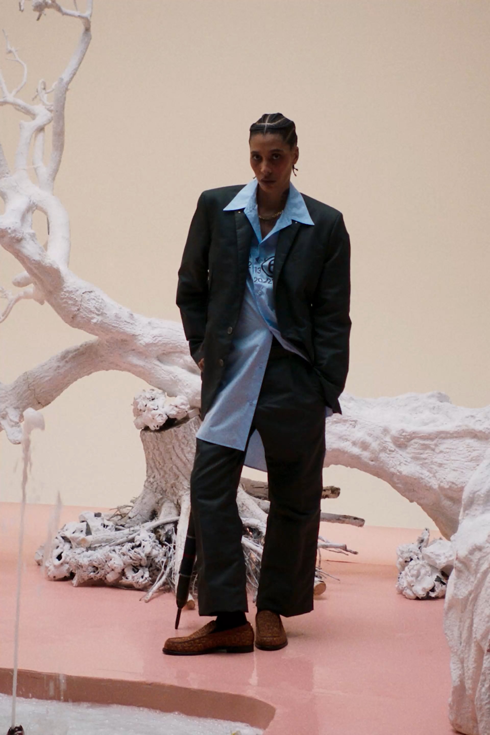 El modelo se presenta con confianza, con las manos en los bolsillos, vestido con un traje negro sobre una camisa azul, al lado de una escultura de árbol blanca sobre un suelo rosa.