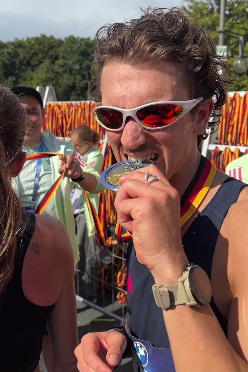 Stříbrná medaile s modrou stuhou držená v ústech. Účastník má na sobě sportovní sluneční brýle a tílko a na zápěstí hodinky.