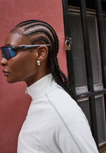 Γυναίκα με κοτσίδες cornrow, φορά μπλε γυαλιά ηλίου και ασημένια σκουλαρίκια κρίκους, ντυμένη με λευκό τοπ με φερμουάρ, μπροστά σε κόκκινο τοίχο.