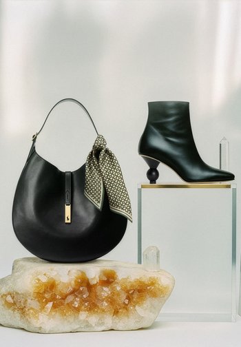 Juodos odos rankinė su raštuoto šalikėlio akcentu ir auksinės spalvos detalėmis šalia juodos odos kulkšnies aukštakulnio su skulptūriniu kulnu.