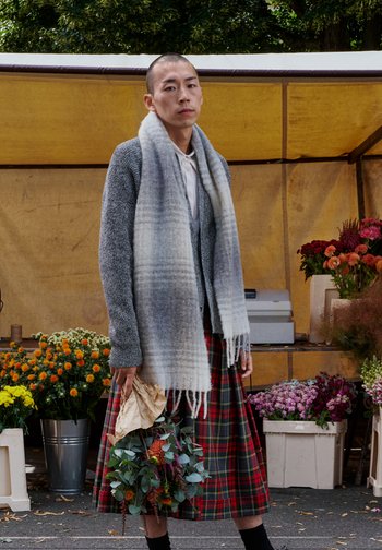 Grijze gebreide cardigan, witte blouse, geruite rok in rood en groen, lichtgrijze sjaal met franjes, met een bloembouquet gewikkeld in papier.