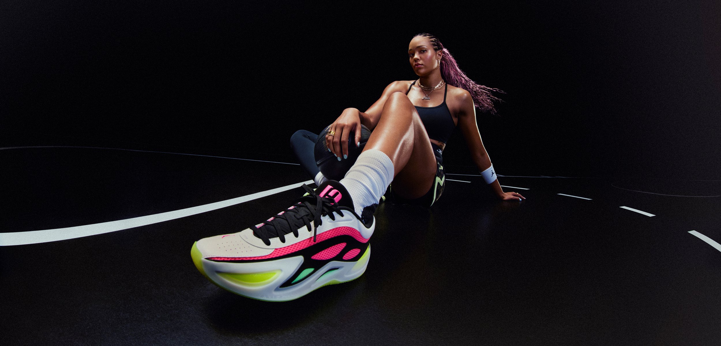 Sportlane spordiriietes istub pimedal väljakul, sirutades ühe jala ette ning esitades neoonaktsentidega jooksujalatseid ja keskendunud ilmet.