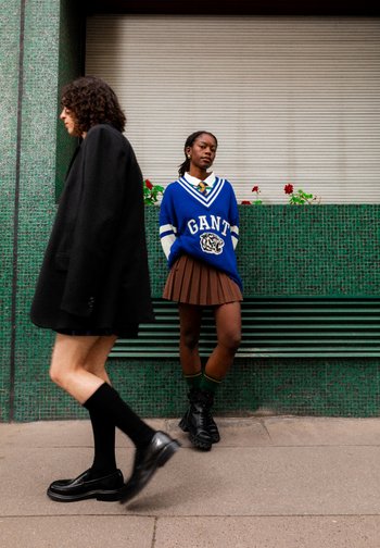 Eine Frau in einem blauen GANT-Pullover und einem braunen Faltenrock steht vor einer grünen Wand, während eine andere Person in einem schwarzen Outfit vorbeigeht.
