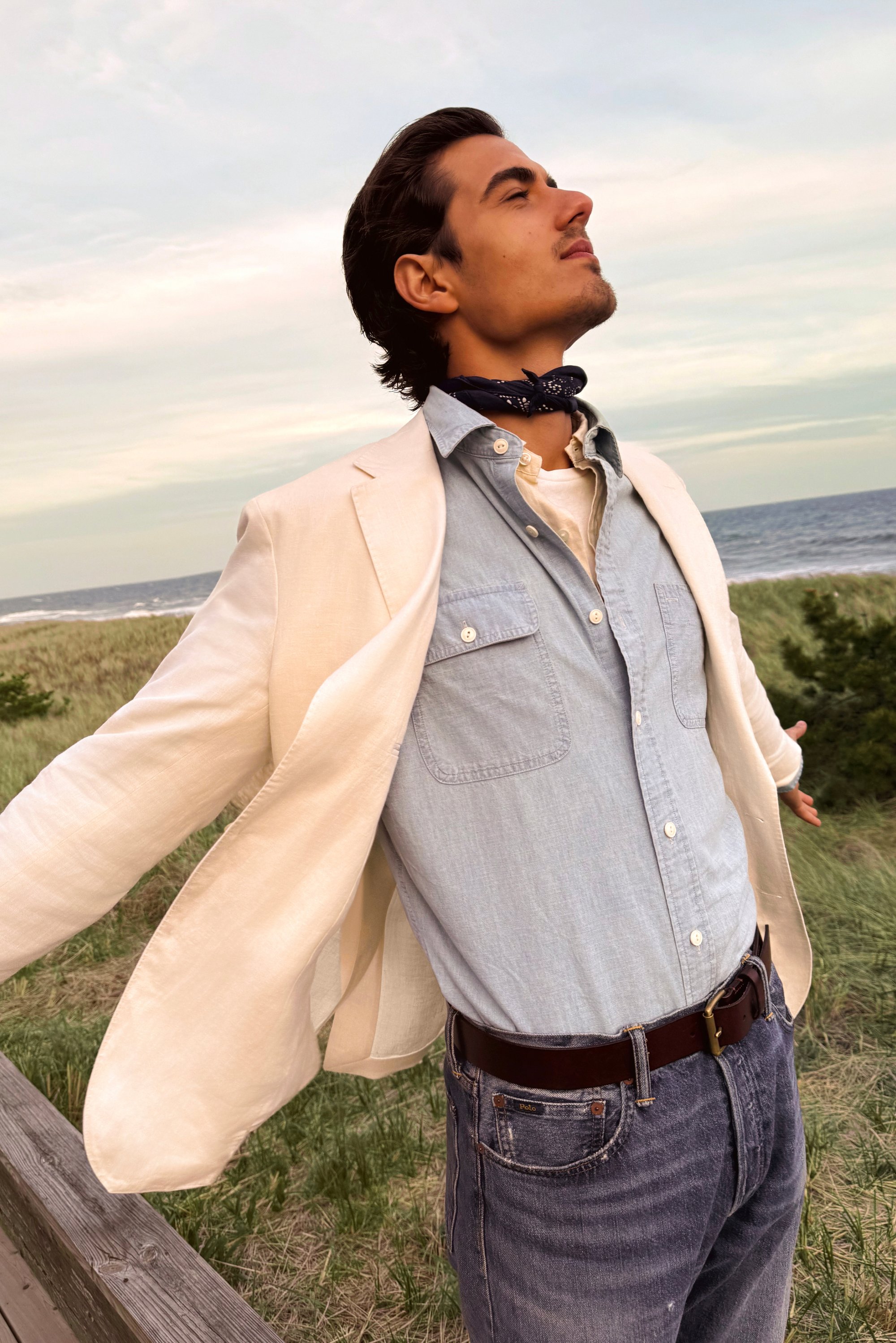 A man stands outdoors, arms open wide, wearing a light blazer, denim shirt, and scarf, with the ocean and grass in the background.