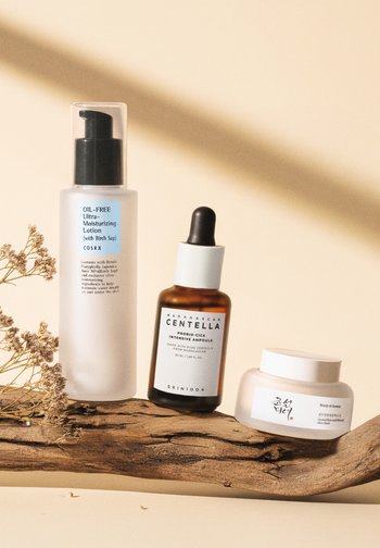 Trois produits de soin de la peau sur du bois : une bouteille à pompe translucide, une bouteille en verre brun avec compte-gouttes, et un pot de crème blanche avec un couvercle en argent.