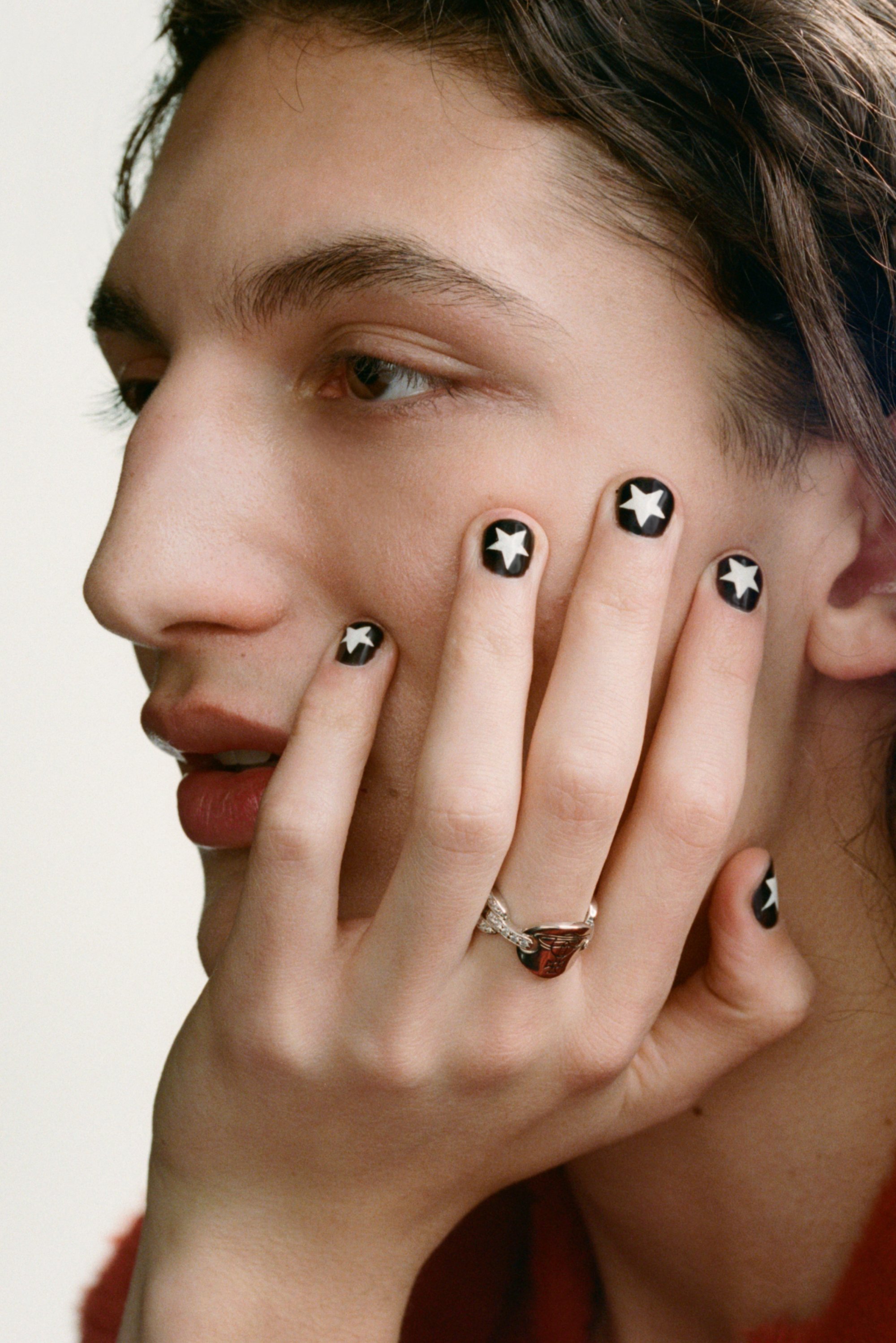 Primer plano de una persona con una expresión neutral, tocándose la cara; uñas pintadas de negro con diseños de estrellas blancas, llevando un anillo ornamentado.