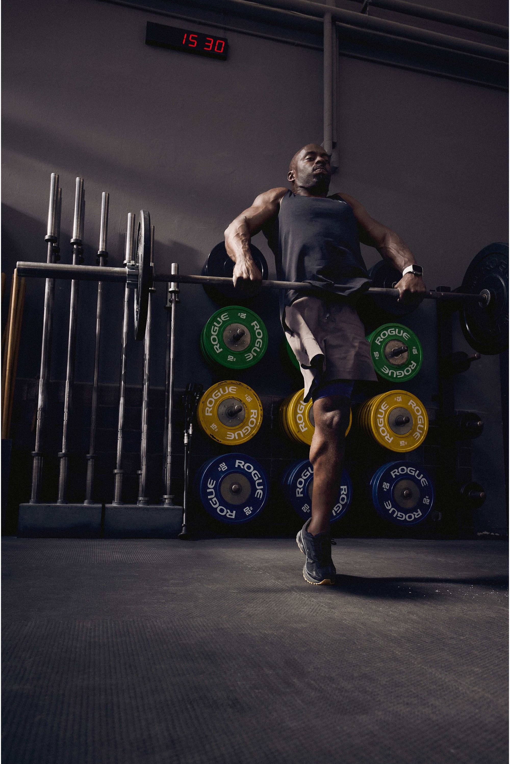 Muž vykonáva cvičenie s činkou v posilňovni, sústredí sa na činku s farebnými závažím naloženými za ním a hodinami ukazujúcimi 15:30.