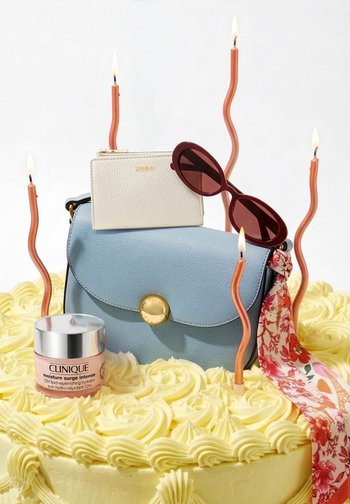 Yellow frosted cake topped with blue handbag, white wallet, red sunglasses, floral scarf, Clinique moisturizer, and five lit wavy candles.