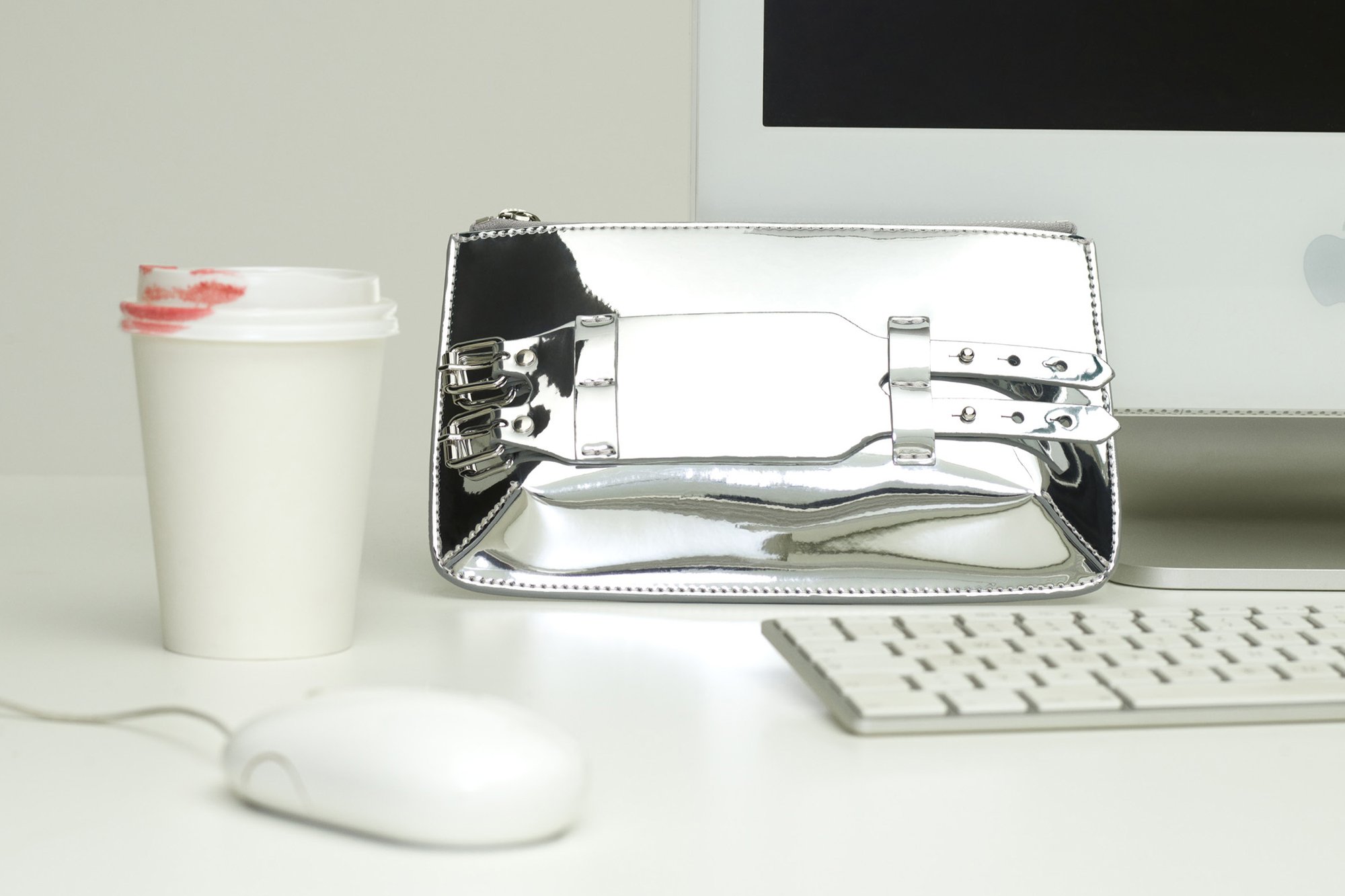 Bolso metálico plateado con un diseño elegante, que cuenta con asas dobles y una textura brillante, colocado al lado de una taza de café blanca y accesorios de computadora.