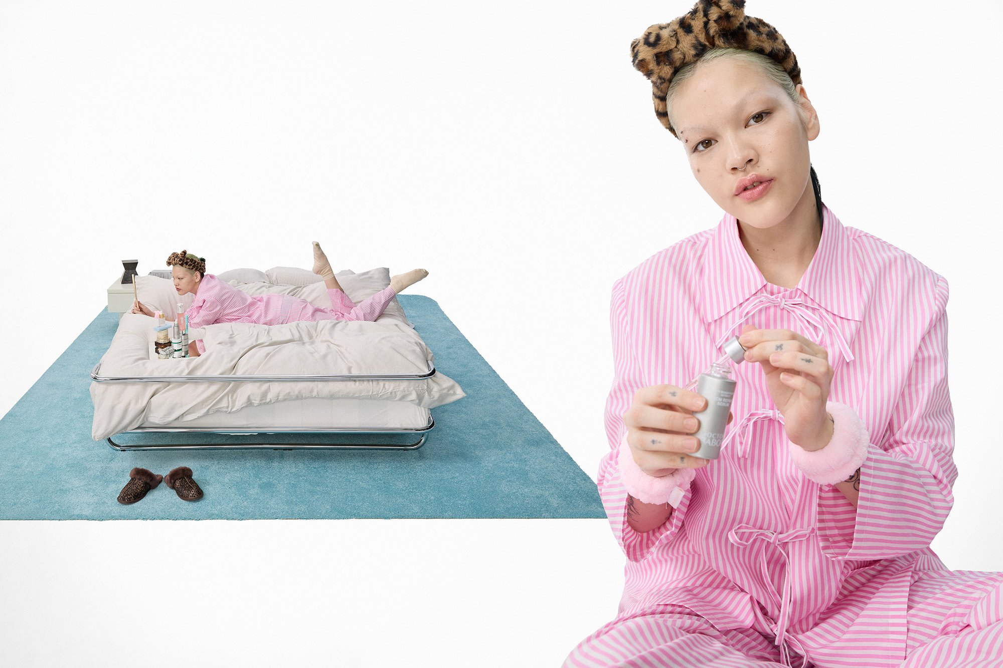 A person in pink striped pajamas and a leopard print headband holds a skincare bottle while lounging on a bed with various products nearby.