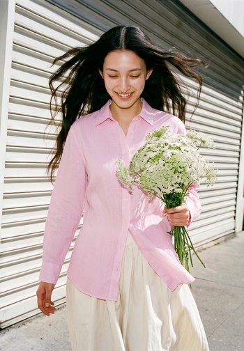 Jeune femme aux longs cheveux souriante tenant un bouquet de fleurs blanches, portant une chemise rose et marchant à l'extérieur près d'un volet métallique.