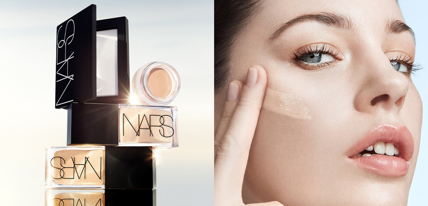 Gleich gestapelte NARS Foundation-Flaschen neben einer Frau, die mit den Fingern cremige Foundation auf ihre Wange aufträgt.