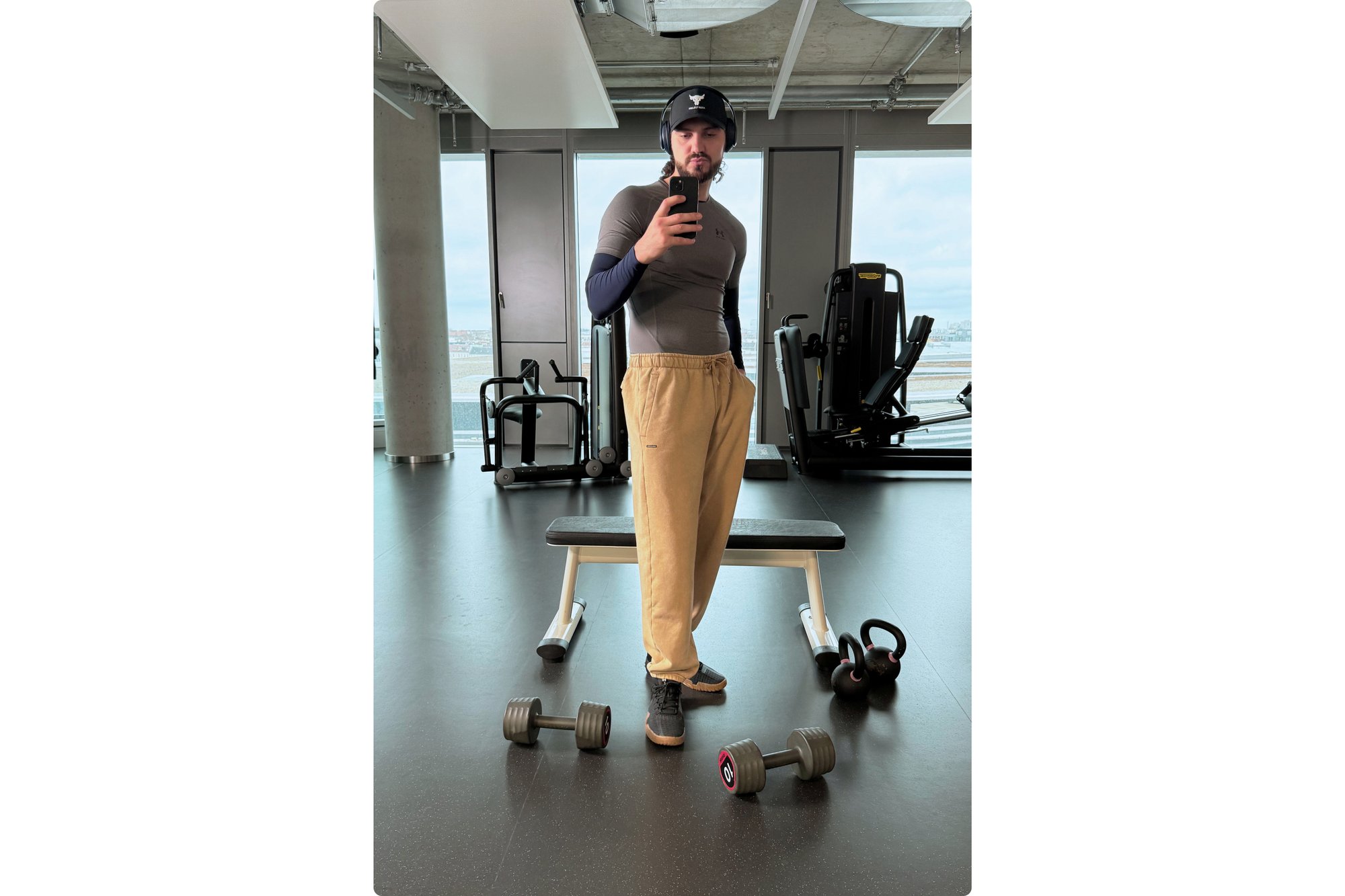 Una persona vestida con una camiseta ajustada y pantalones de chándal se encuentra en un gimnasio, sosteniendo un teléfono, con mancuernas y un banco de ejercicios cerca.